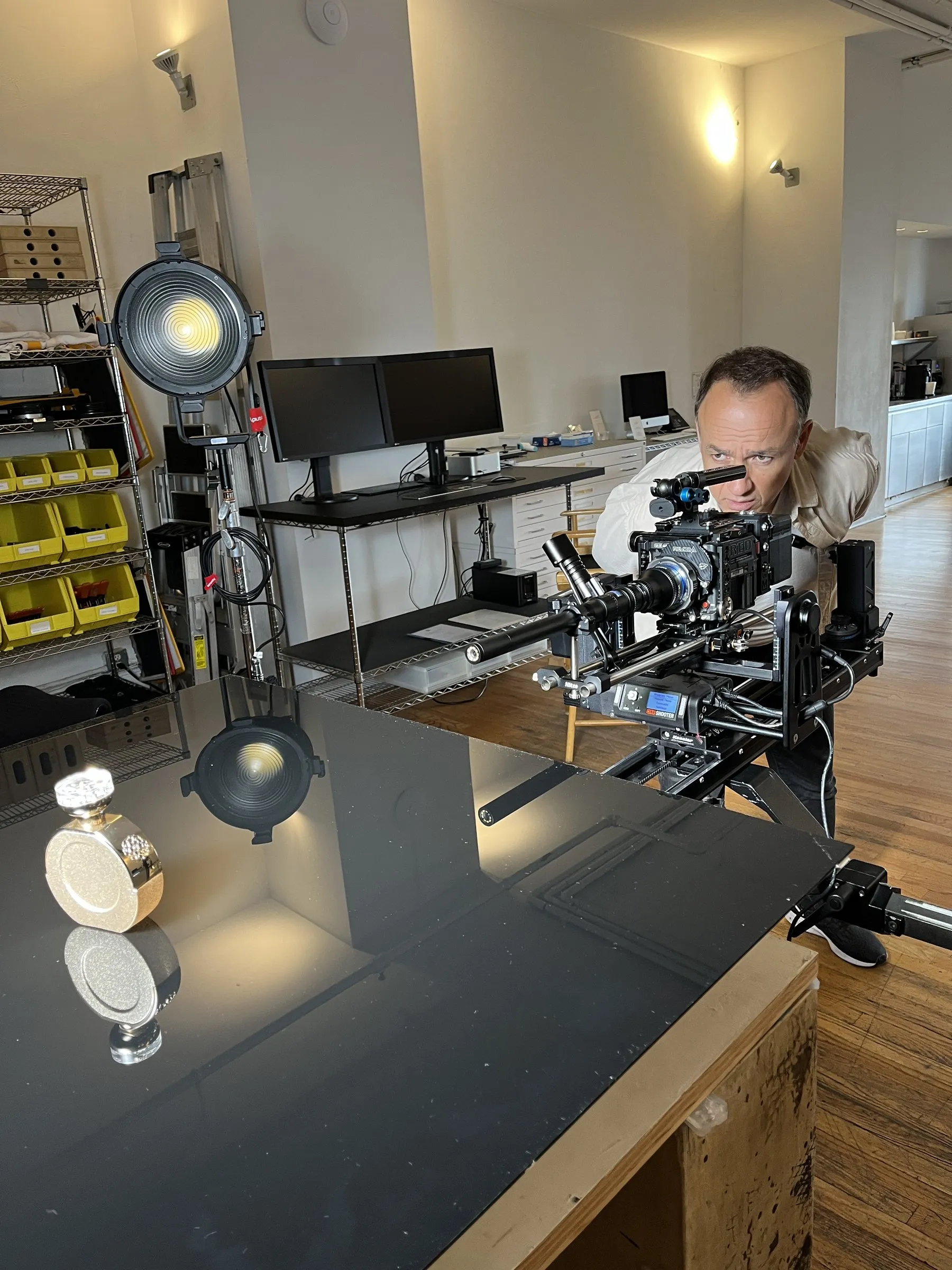 A cinema camera on a rig at a tabletop set, the operator at the eyepiece, a small product under the light.